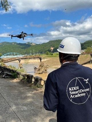 写真：川沿いでドローンを操縦する人と飛行するドローン