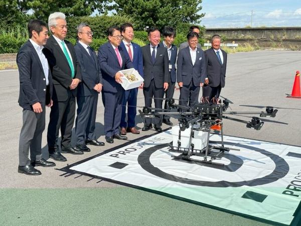 写真：ドローンを前に、横並びに整列する人々。箱を手に持ち中身を見せる大村知事。