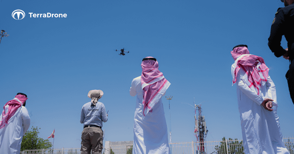 写真:操縦者など複数の人の向こうで飛行するドローン