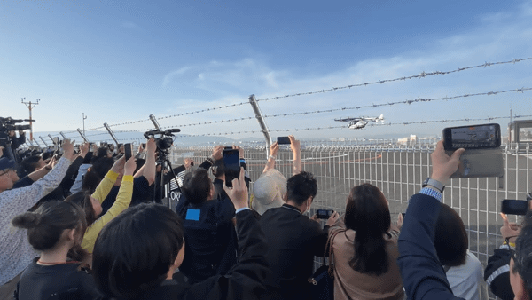 写真：フェンス越しに飛行する空飛ぶクルマ、その様子をスマートフォンで撮影する大勢の人