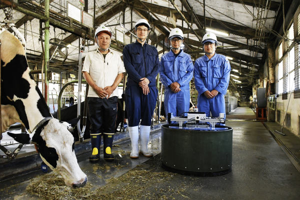 写真：牛舎内の餌寄せロボットと並ぶ4人、首を伸ばしてわらを食べる牛