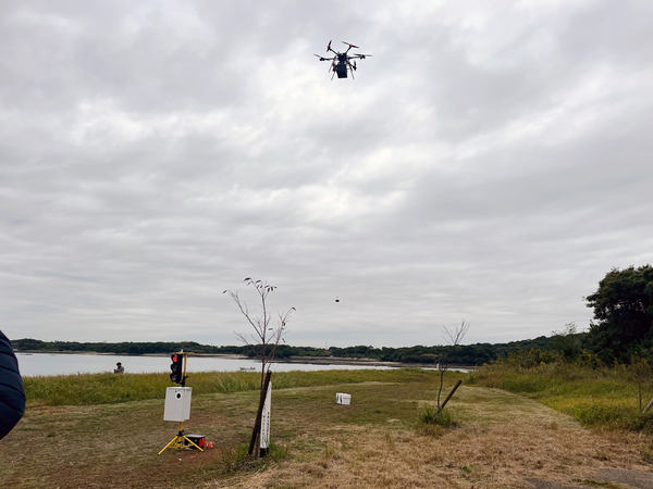 写真：河原に設置された簡易的な信号（赤）、その向こう側で上空から荷物を下ろすドローン