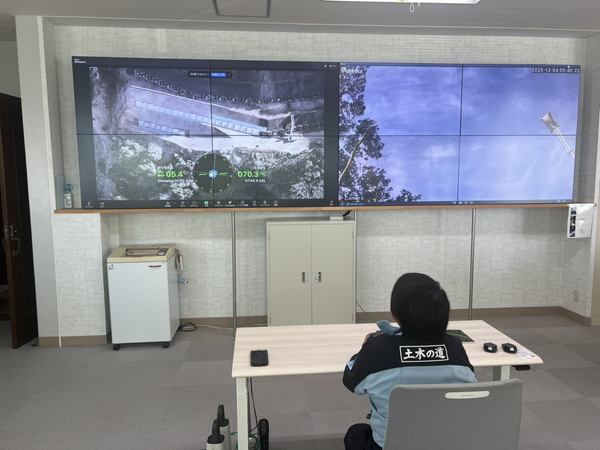 写真:屋内のディスプレイに映ったドローンのカメラ映像と俯瞰カメラ映像、その映像を確認しながら操縦するスタッフ