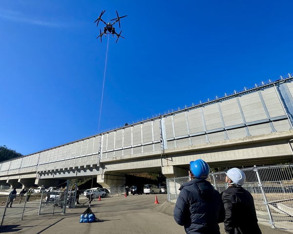 写真：荷物を吊り下げたドローンが飛行する様子、それを見上げる作業員